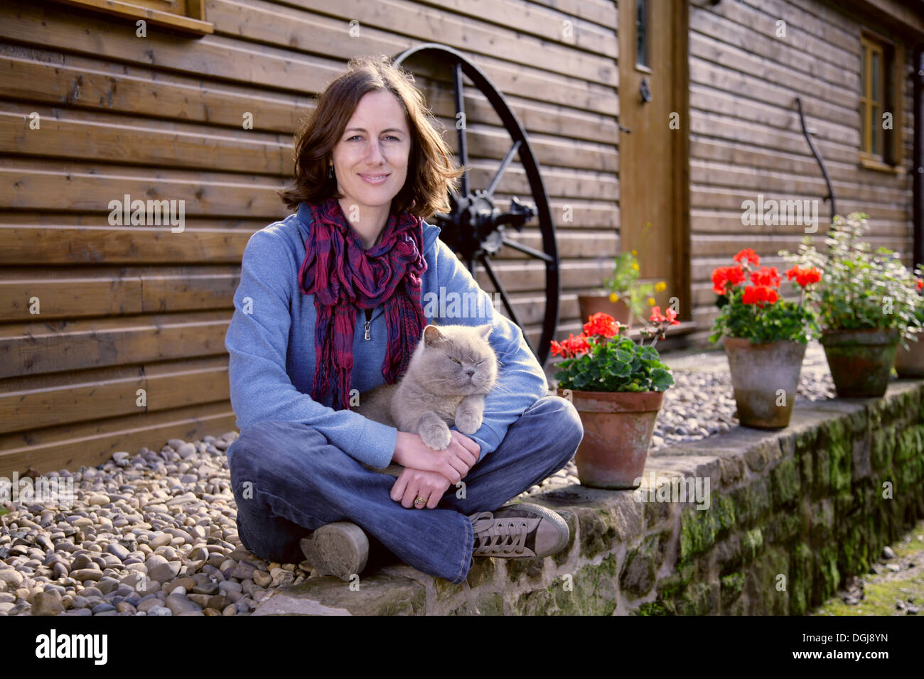 Junge Frau mit einer grauen Katze auf dem Schoß auf einer Mauer sitzend. Stockfoto