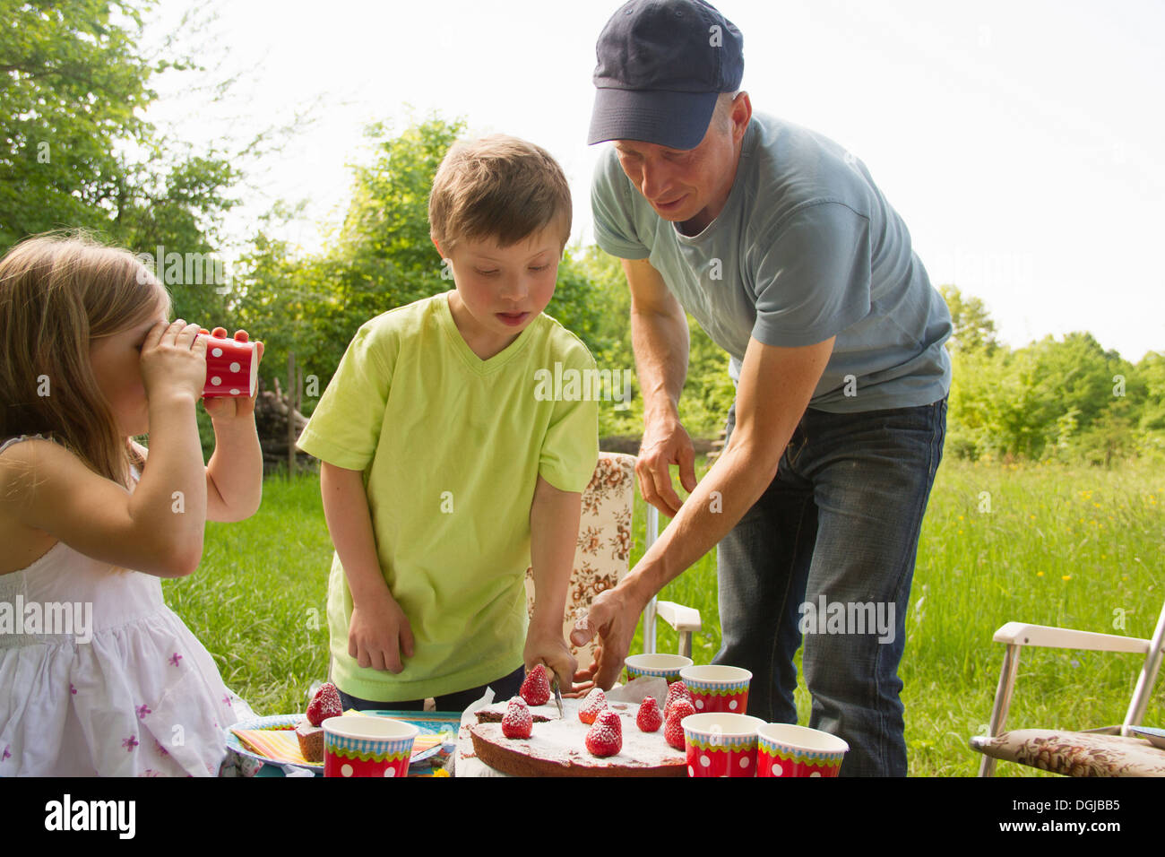 Vater mit zwei Kindern schneiden Geburtstagskuchen im freien Stockfoto