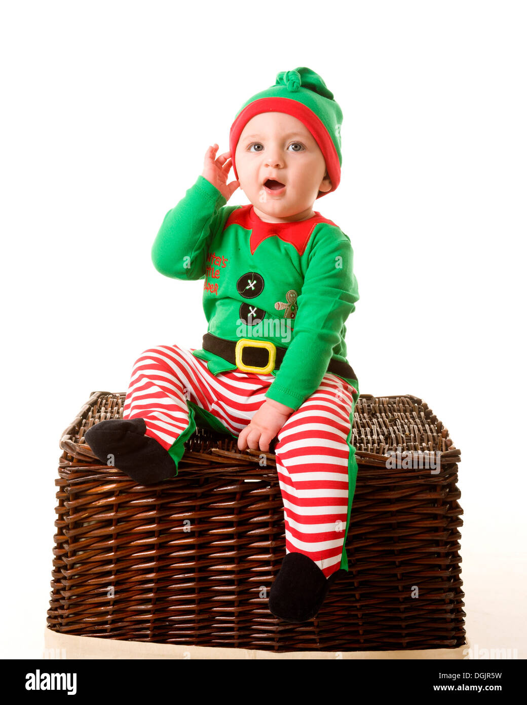 Ein Kind, das wie eine Weihnachtselfe gekleidet ist und auf einem Korbkorb sitzt, mit der Hand zu Ohren sitzt, als ob es auf den Weihnachtsmann hört Stockfoto
