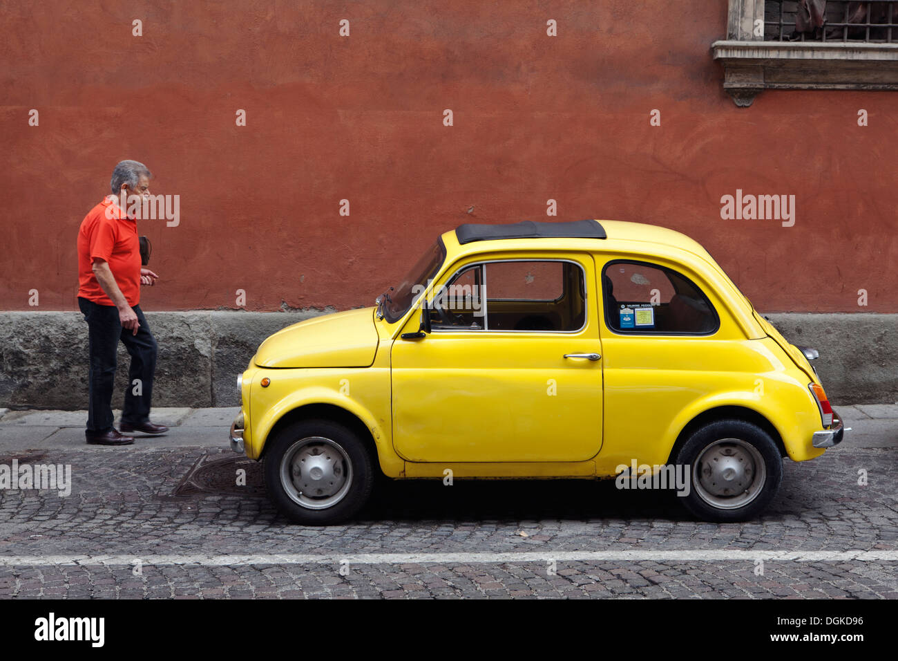 Ein Mann geht über einem gelben Fiat 500 Oldtimer Parken auf einer gepflasterten Straße vor einem ockerfarbenen Wand in Bologna. Stockfoto