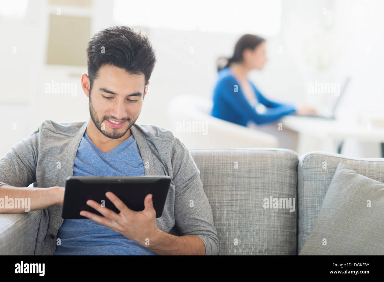 Mann, sitzend auf Sofa mit digitalen Tablet zu Hause, Frau im Hintergrund Stockfoto