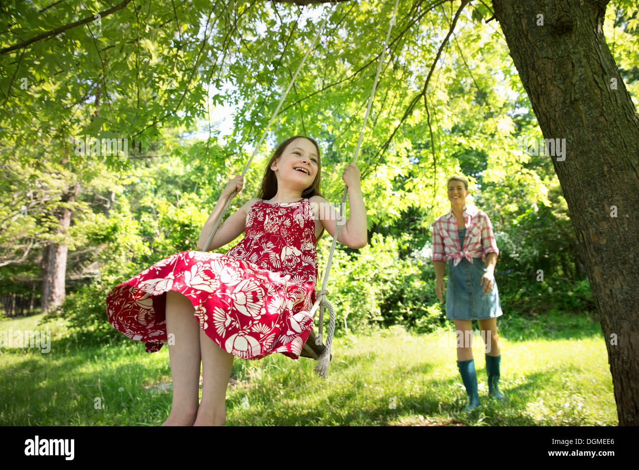 Sommer. Ein Mädchen in einem Sommerkleid auf einer Schaukel unter einem grünen Baum. Eine Frau, die hinter ihr stehen. Stockfoto