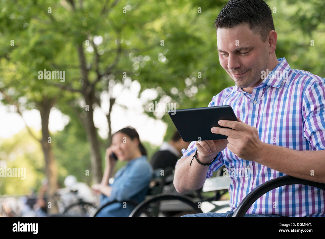 Sommer. Eine Frau und zwei Männer sitzen im Park, jede auf ihrem eigenen Handy oder verwenden eines Tablet. Stockfoto