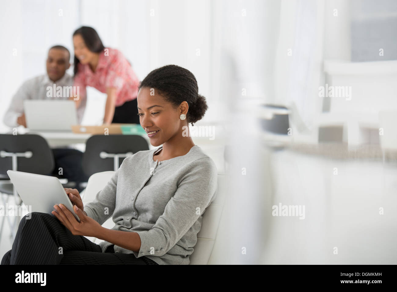 Geschäft. Eine Frau mit einem digitalen Tablet sitzen. Zwei Personen im Hintergrund, einem Computer-Bildschirm betrachten. Stockfoto