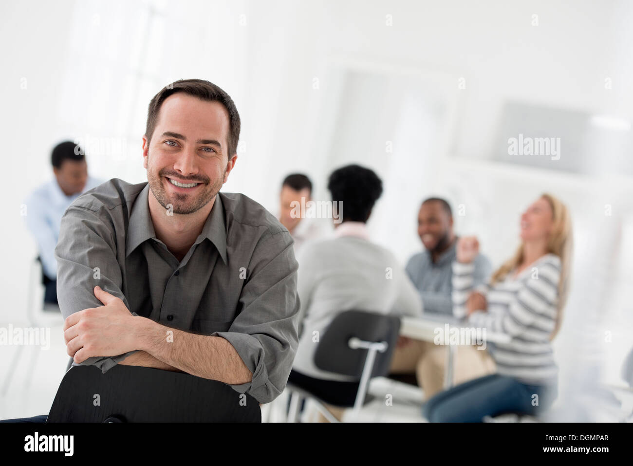 Büroeinrichtung. Ein Mann sitzt mit seinen bewaffneten gefaltet, Blick in die Kamera. Eine Gruppe von Menschen an einem Tisch im Hintergrund. Stockfoto