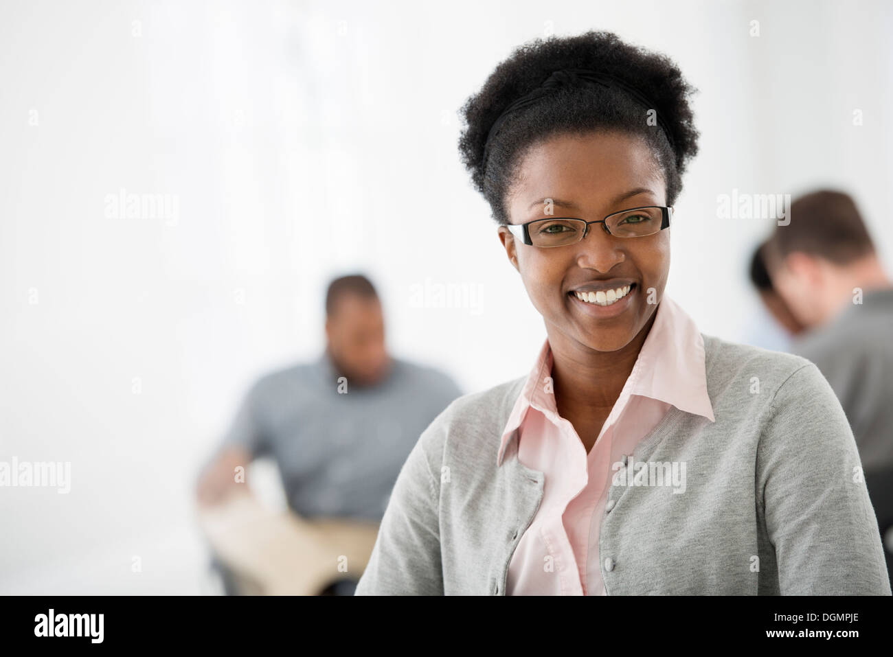 Büroeinrichtung. Eine Frau lächelt selbstbewusst. Zwei Kollegen im Hintergrund. Stockfoto
