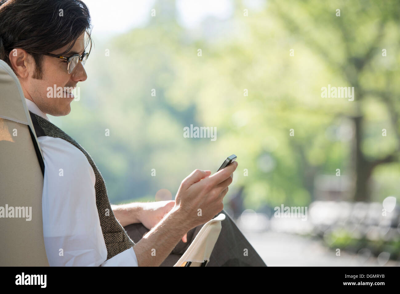Leben in der Stadt. Ein Mann sitzt in einer Leinwand Campingstuhl in den Park, Blick auf sein Handy. Stockfoto