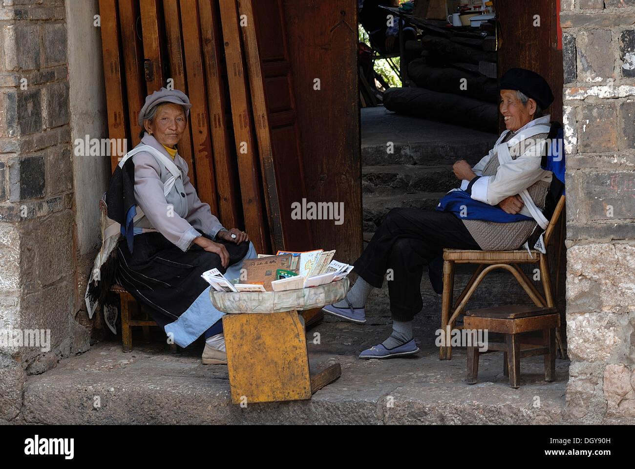 Zwei Frauen in der Tracht der Naxi-Minderheit sitzen mit einem kleinen Verkaufsstand in einem Hauseingang, Lijiang, Yunnan Stockfoto