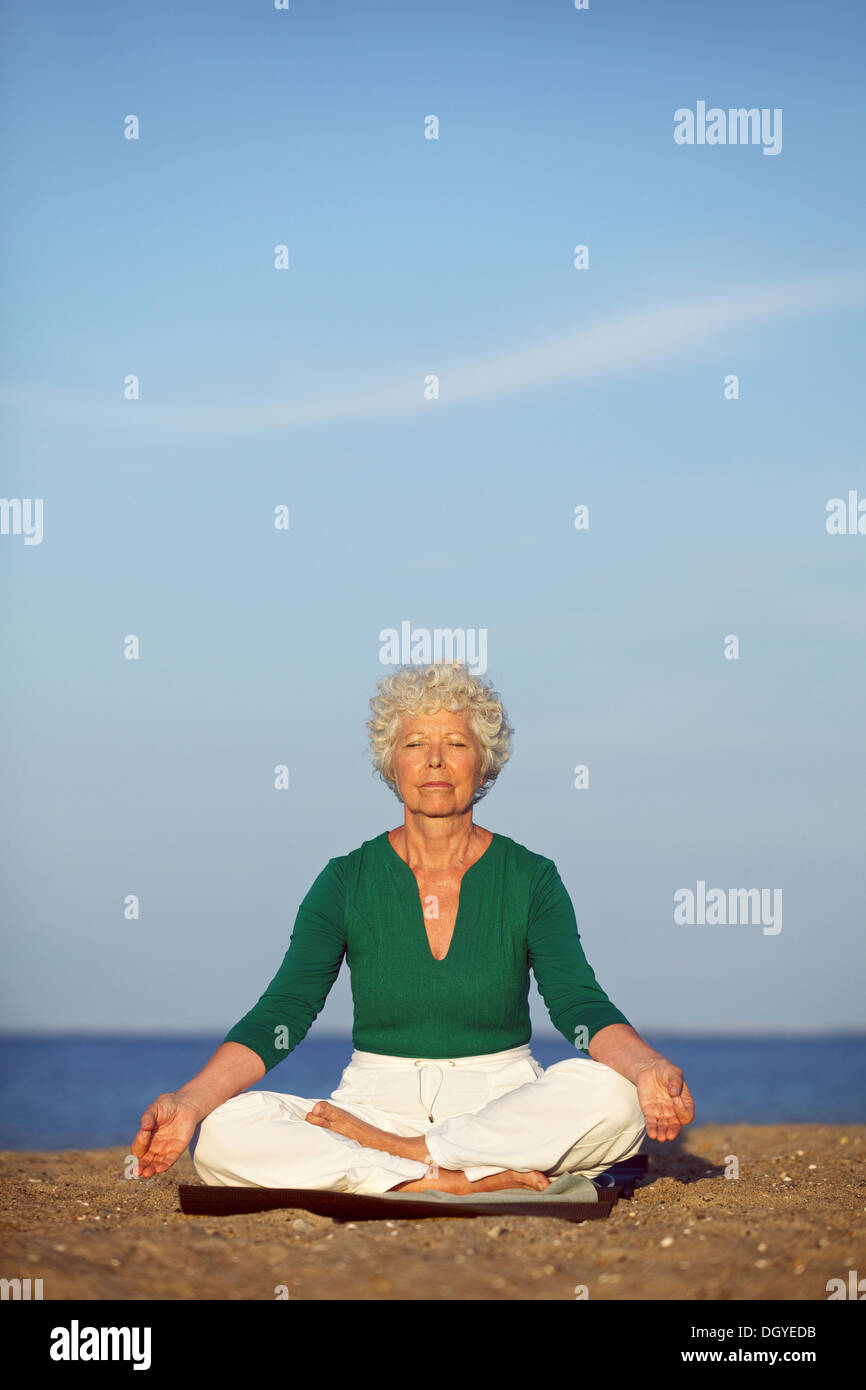 Ältere Frau praktizieren Yoga am Morgen mit Meer im Hintergrund. Alten kaukasischen Frau Entspannungsübung am Strand zu tun. Stockfoto