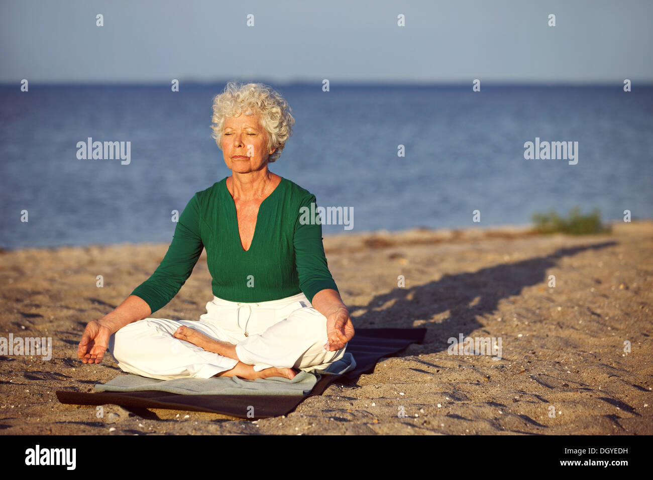 Ältere Frau sitzen auf Gymnastikmatte Meditation in Lotus-Pose am Strand zu tun. Alte Frau Entspannungsübung zu tun Stockfoto