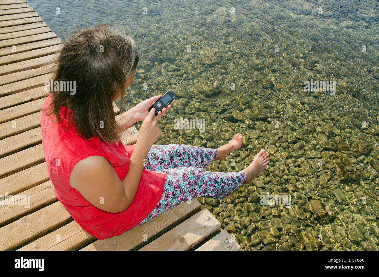 Frau sitzt auf einem Dock am Wasser mit einem Handy, Tisno, Murter Insel, Adria, Dalmatien, Kroatien, Europa Stockfoto