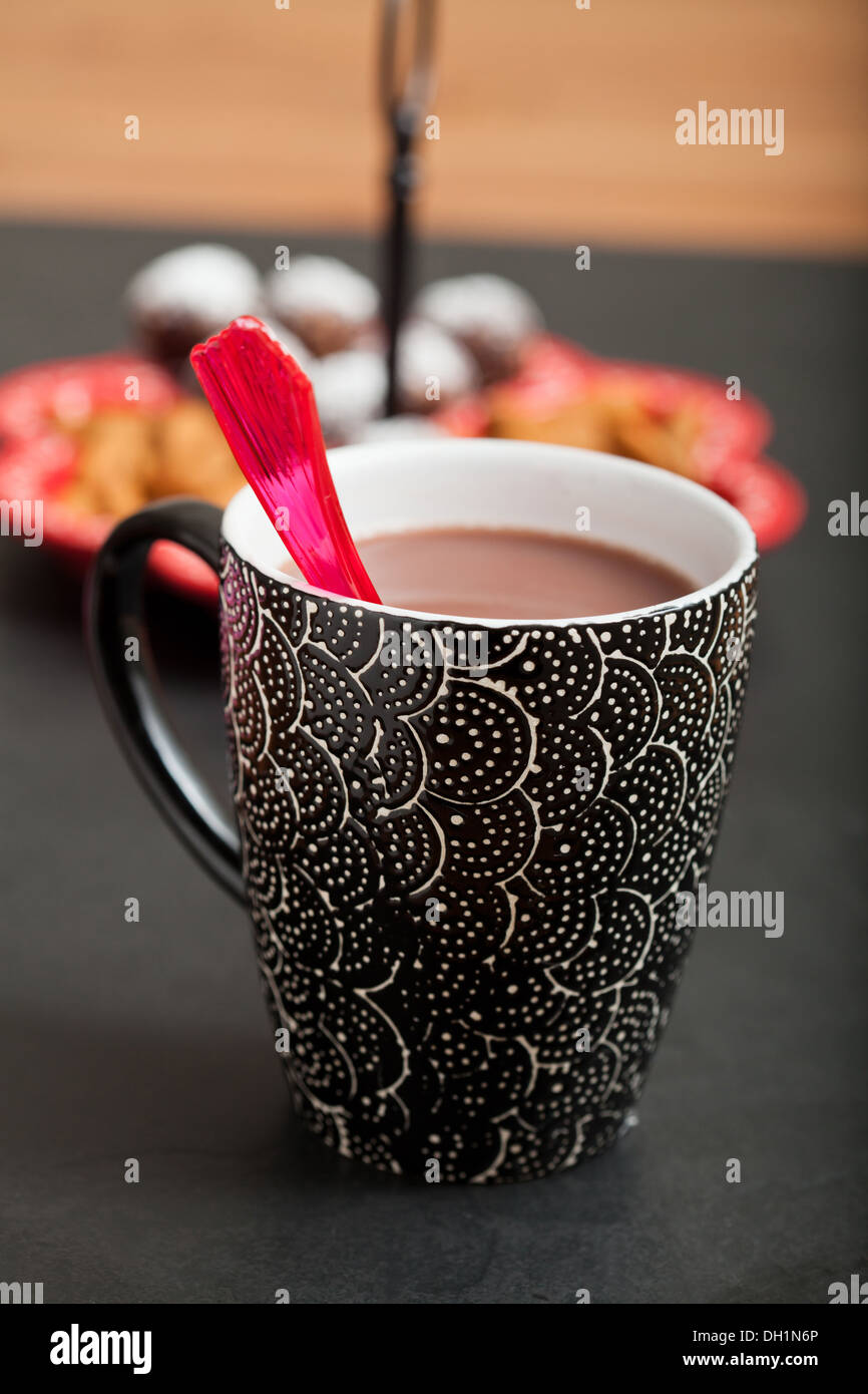 Nahaufnahme der Tasse heiße Schokolade mit roten Plastiklöffel mit Tablett mit Schokokugeln und Mandeln im Hintergrund Stockfoto