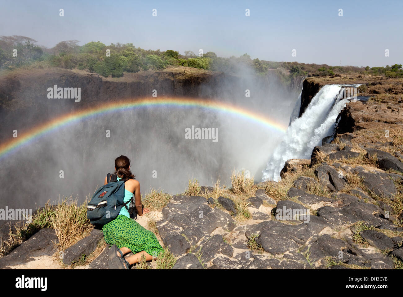 Alleinreise; EINE alleinstehende Frau, die auf der Seite von Livingstone Island, Sambia Africa, den Regenbogen über den Victoriafällen betrachtet Stockfoto