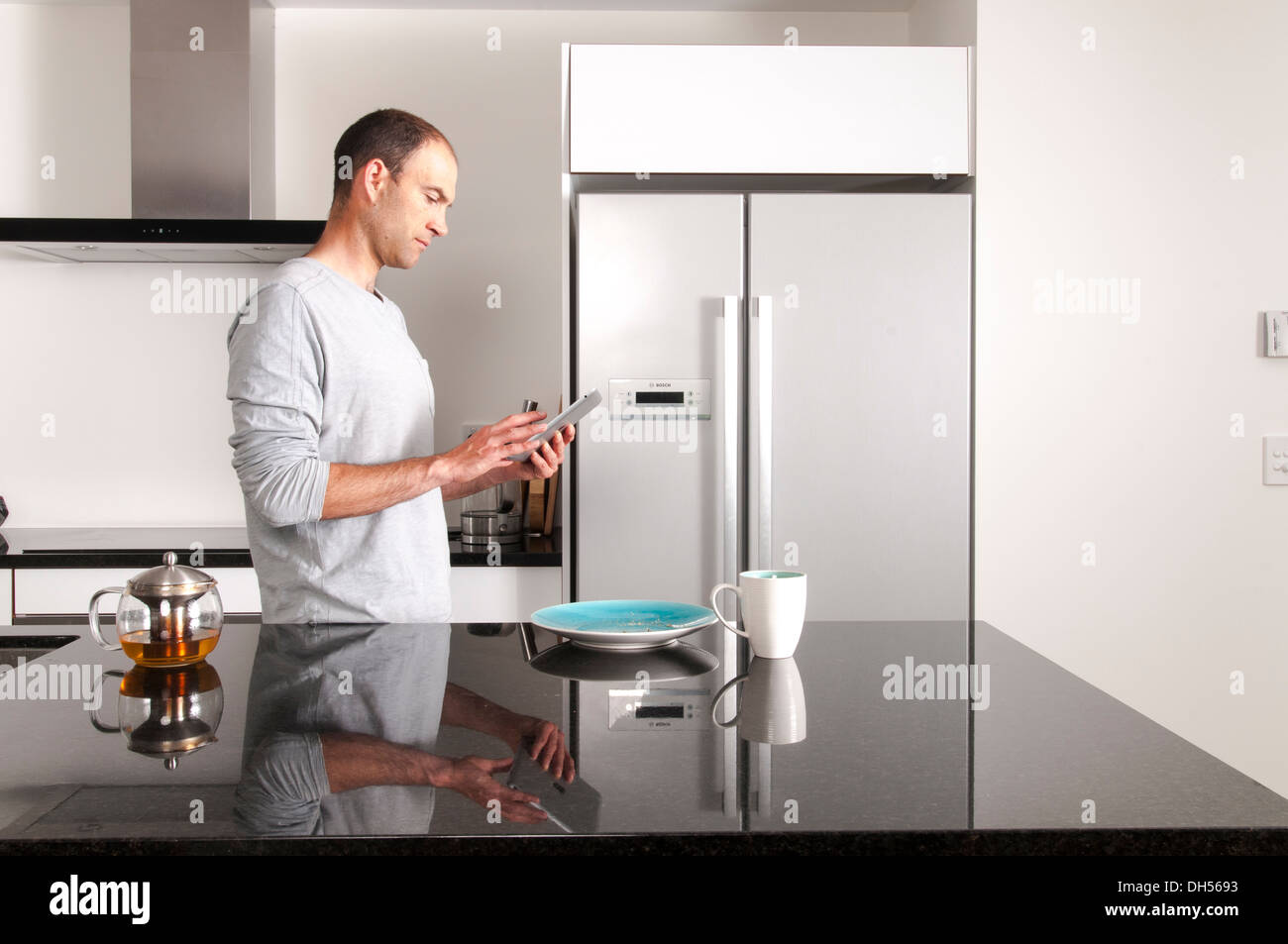 Mann hält Ipad in modernen Küche mit Tasse grüner Tee und Platte auf Granit-Arbeitsplatte. Stockfoto