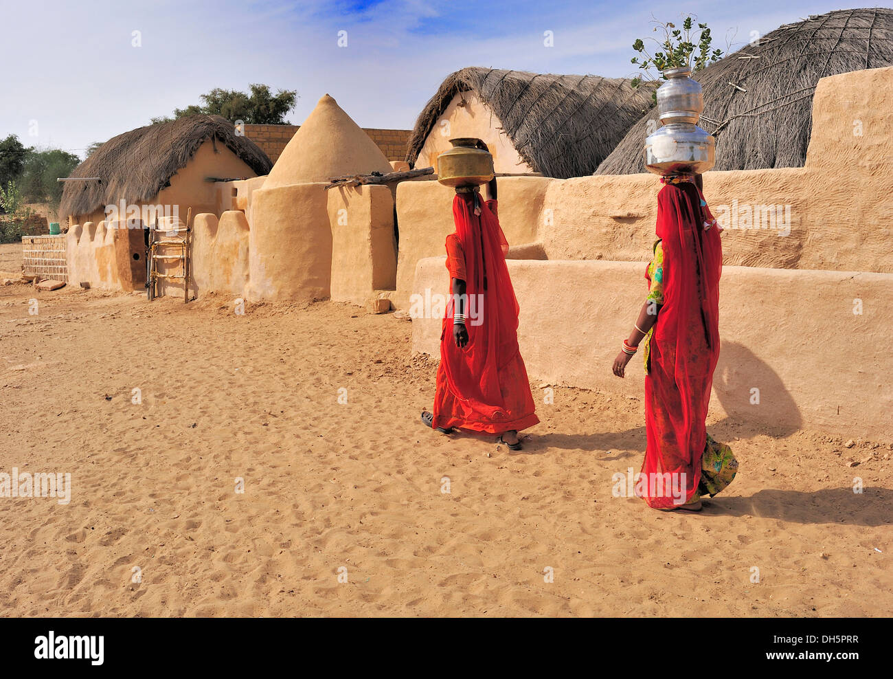Zwei indische Frauen tragen rote Saris tragen von Wasser gefüllten Krügen auf dem Kopf durch ein Dorf, Thar-Wüste, Rajasthan, Indien Stockfoto