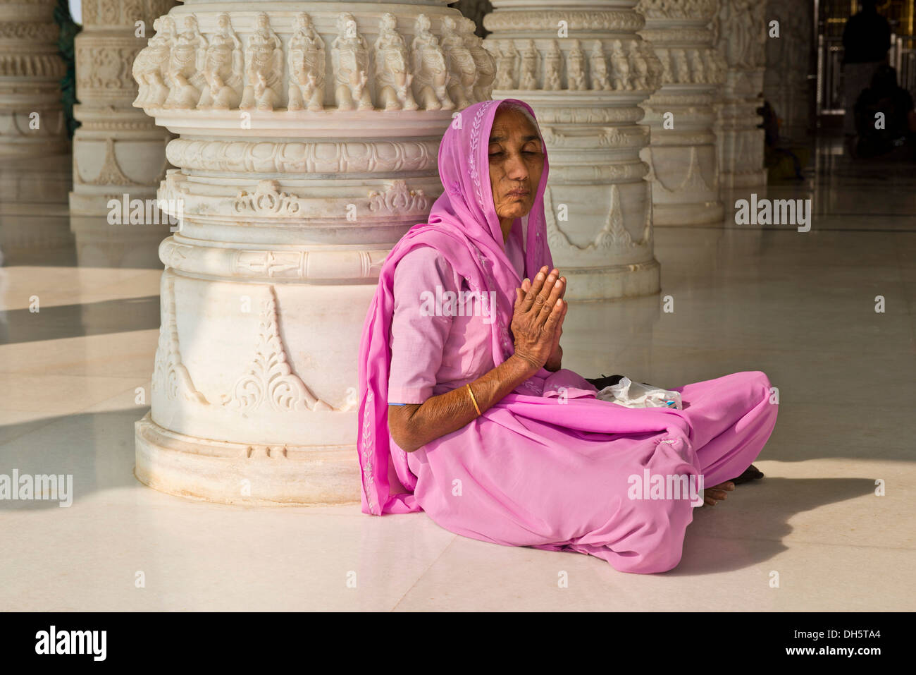 Alte indische Frau in einen Sari sitzen auf dem Boden im Gebet, vor einem reich verzierten Marmorsäule am Shri Swaminarayan Mandir, ein Stockfoto