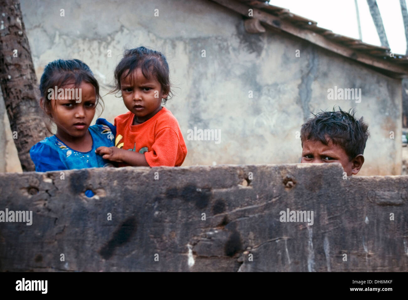 drei kleine Kinder stehen hinter einer Mauer außerhalb ihrer verarmten Heimat in Benaulim Goa Indien Stockfoto