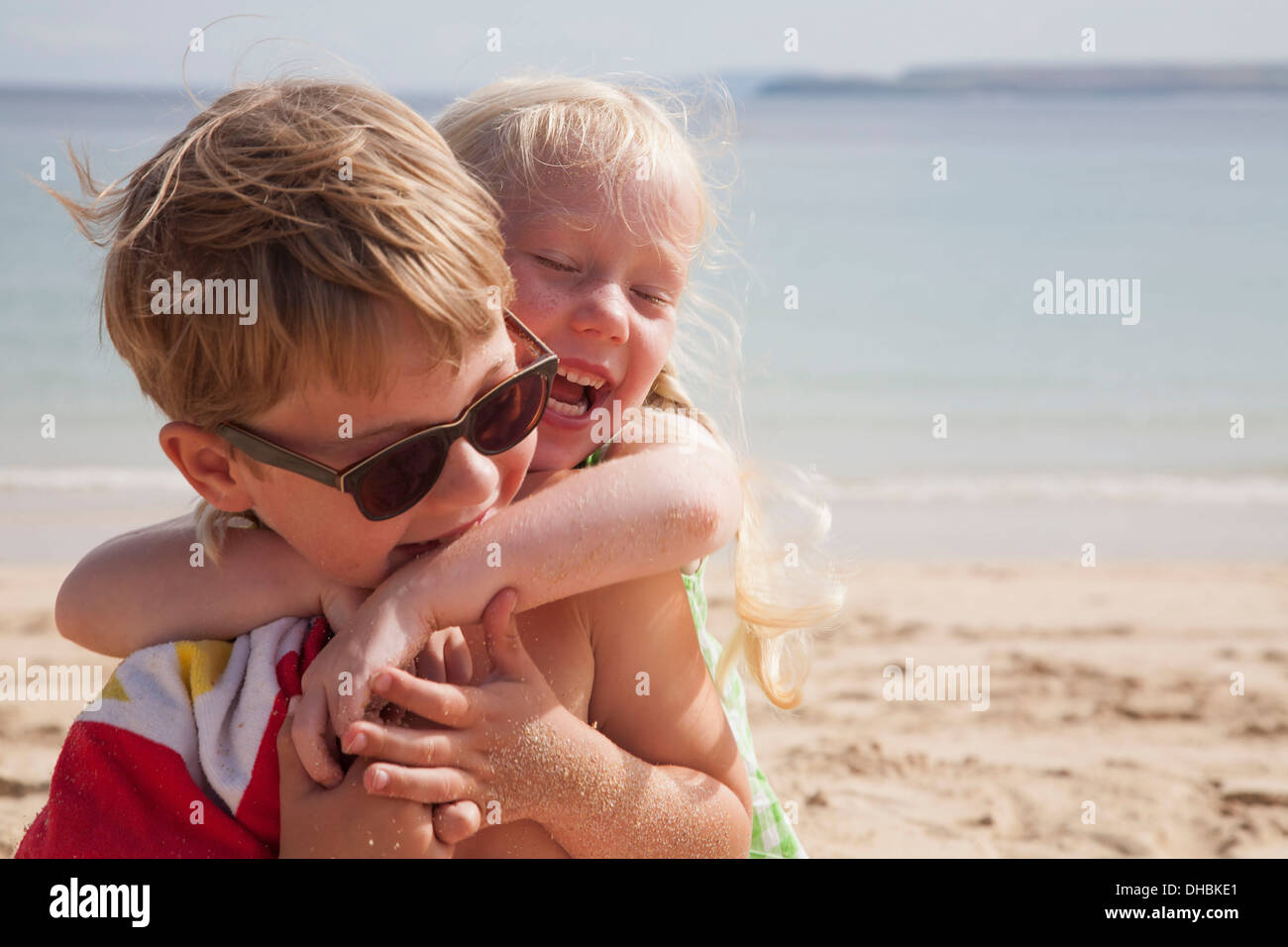 Bruder und Schwester spielen kämpfen am Strand. Ein Junge in einer Sonnenbrille und einem jüngeren Mädchen mit ihre Arme um seinen Hals. Stockfoto