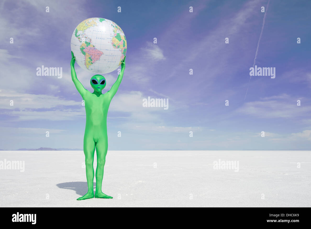 Grüne Alien hält einen Globus der Erde über seinem Kopf stehend auf einsamen weißen Wüstenplaneten Stockfoto