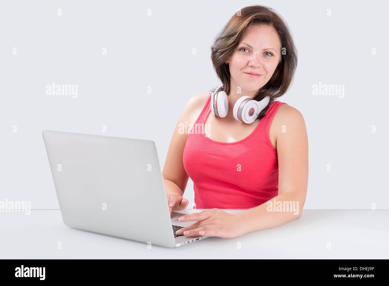 Junge Frau sitzt vor Laptop mit Bluetooth Kopfhörer an ihrem Hals und schaut in die Kamera Stockfoto