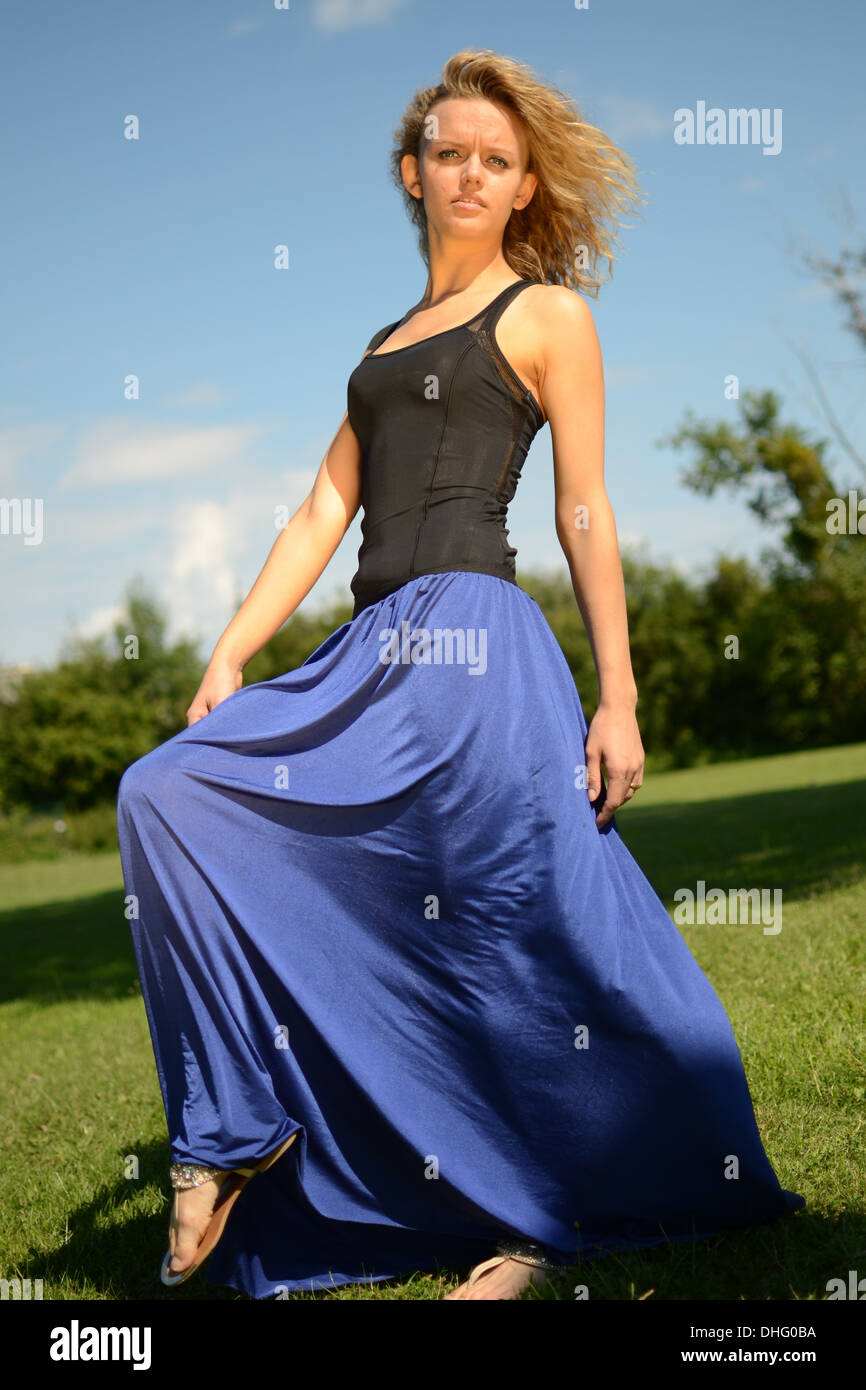 Weibliches Model mit blonden, lockigen Haaren. Mädchen mit blauen Kleid und schwarzen Top. Outdoor-Foto-Session im Park mit blauem Himmel. Stockfoto