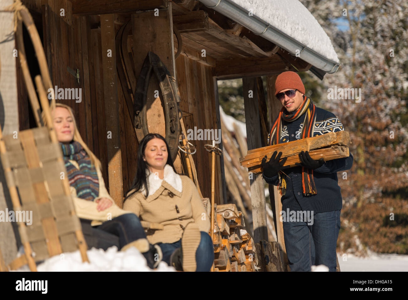Junger Mann tragen Holzscheit Winter Hütte Schnee Freundinnen Sonnenbaden Stockfoto