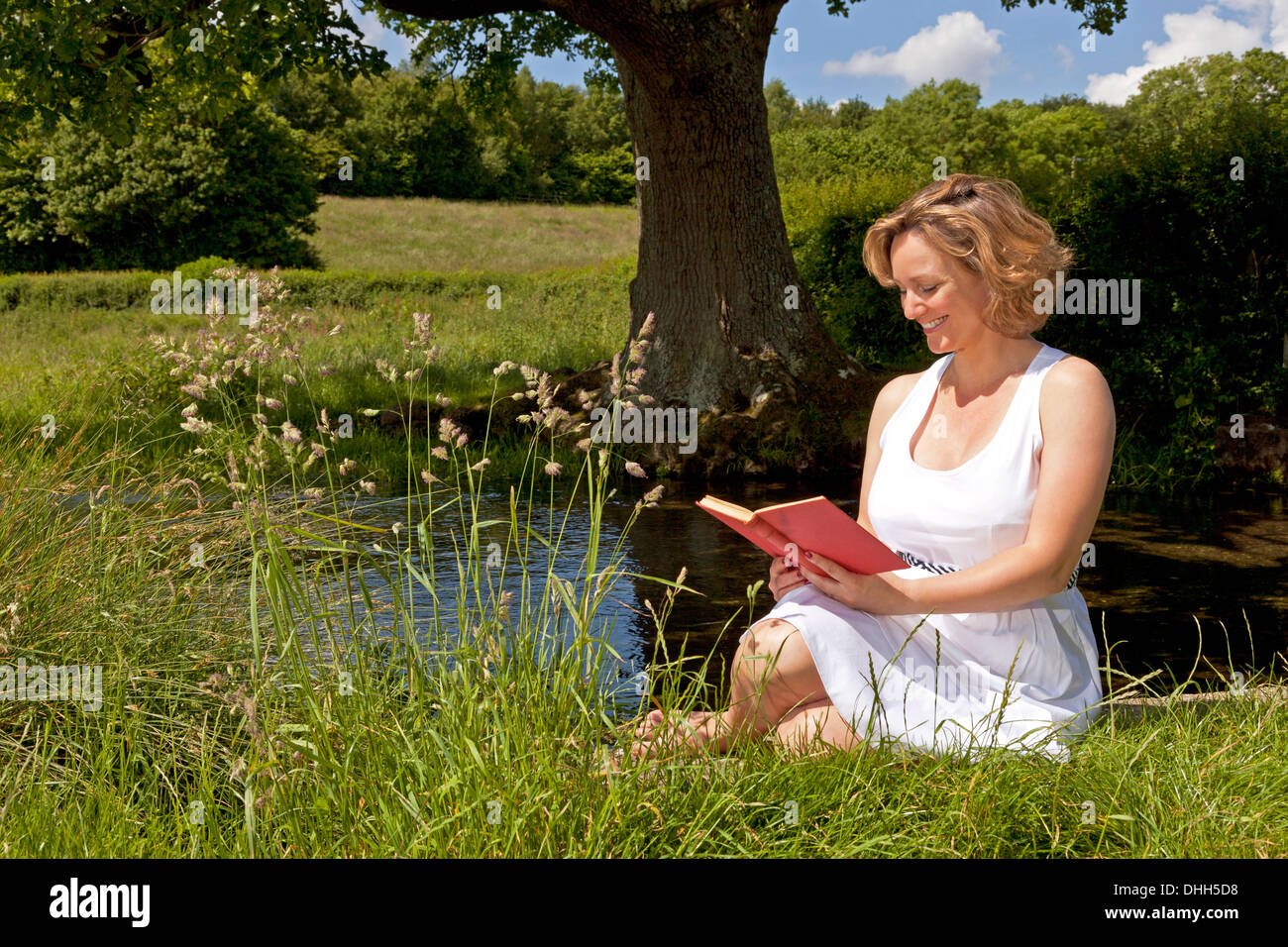 Eine Frau in einem weißen Kleid saß an einem Bach, ein Buch zu lesen, an einem hellen Sommertag. Stockfoto