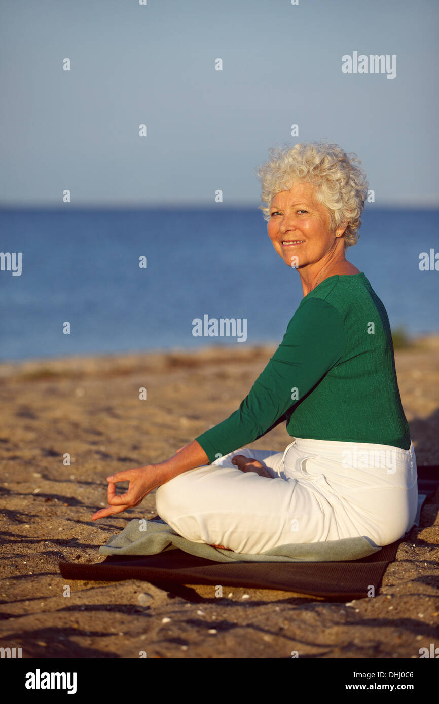 Glückliche alte Frau gehen Entspannungsübung sitzend am Sandstrand. Senior kaukasischen praktizieren Yoga am Meer Morgen Stockfoto