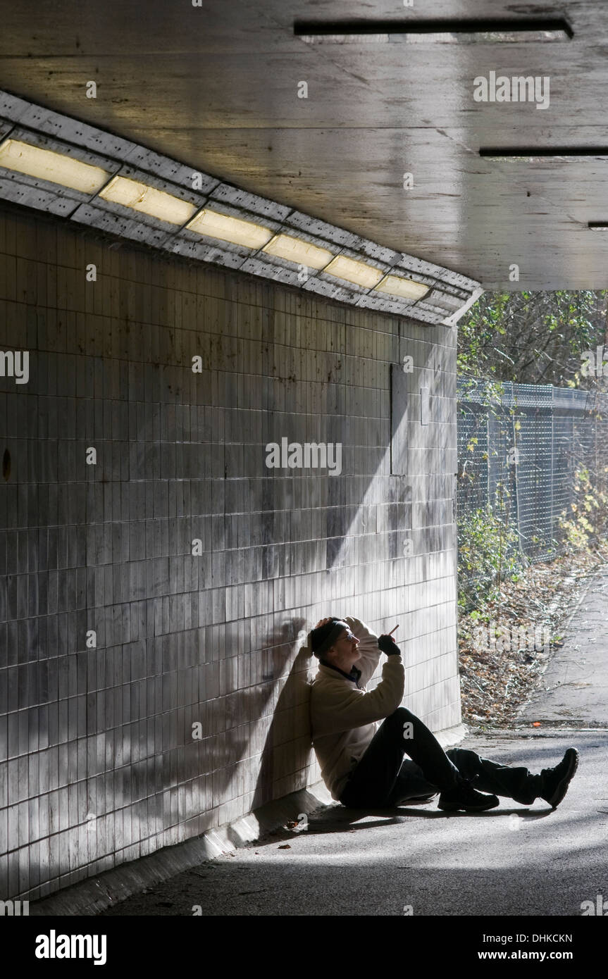 Ein eingefliester Mann, der raucht, sitzt im Sonnenlicht an einer Unterführung und lehnt sich an eine geflieste Wand, um zu kontemplieren. Stockfoto