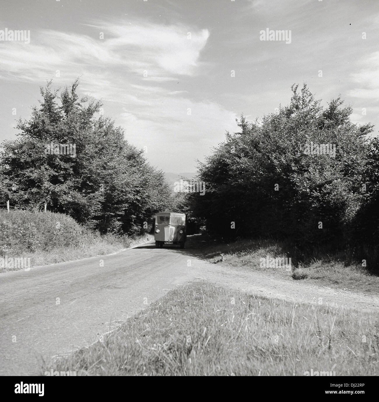 1950er Jahren und ein historisches Bild von van aus den Bäumen auf einer kleinen, leeren Landschaft Straße ohne Markierungen. Stockfoto