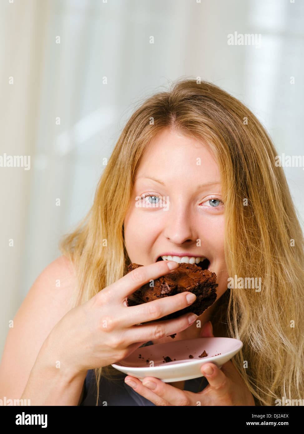 Foto von einer schönen blonden Frau in ihren frühen Dreißigern mit Log blonden Haaren ein großes Stück Brownie oder Kuchen essen. Stockfoto