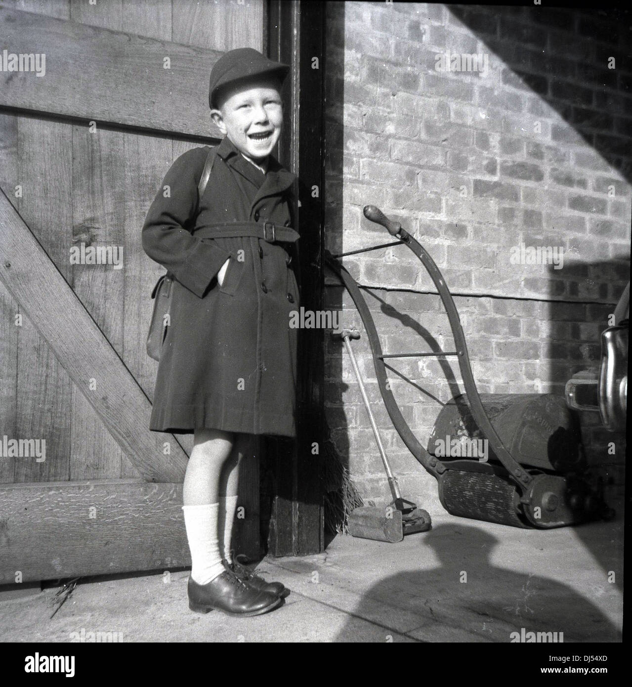 1950s, historisches Bild, ein glücklicher Schuljunge mit Mantel, Mütze und Tasche, der vor einer offenen Tür zu einer Garage mit einem Rasenmäher der Ära im Inneren stand. Stockfoto
