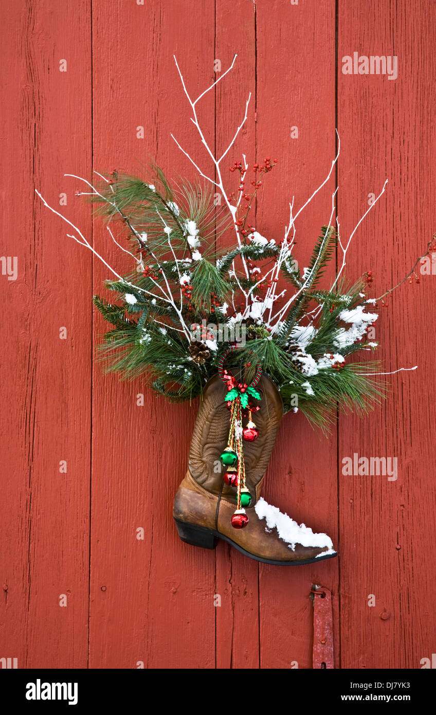 Weihnachtskranz an der Tür, Ranch-Cowboystiefel mit Glocken und immergrünen Ästen an einer alten roten Scheunentür in New Jersey, USA, US-Winter-Schneehumor Stockfoto