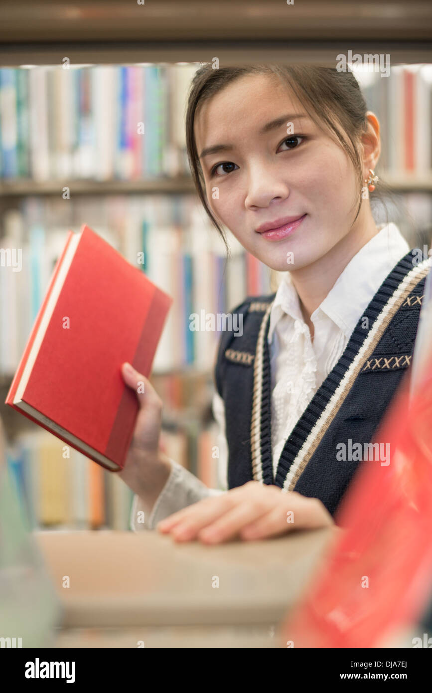 Hübsches junges Mädchen suchte er sich ein rotes Buch aus einem Stapel Bücher auf einem Regal in der Bibliothek Stockfoto