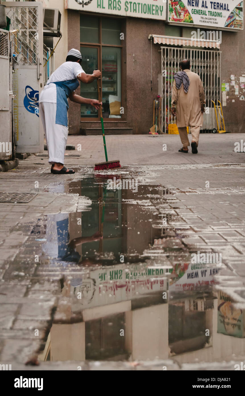 Ein Sachbearbeiter fegt die nassen Bürgersteig vor seinem Laden in den Straßen von Deira. Dubai, Vereinigte Arabische Emirate. Stockfoto