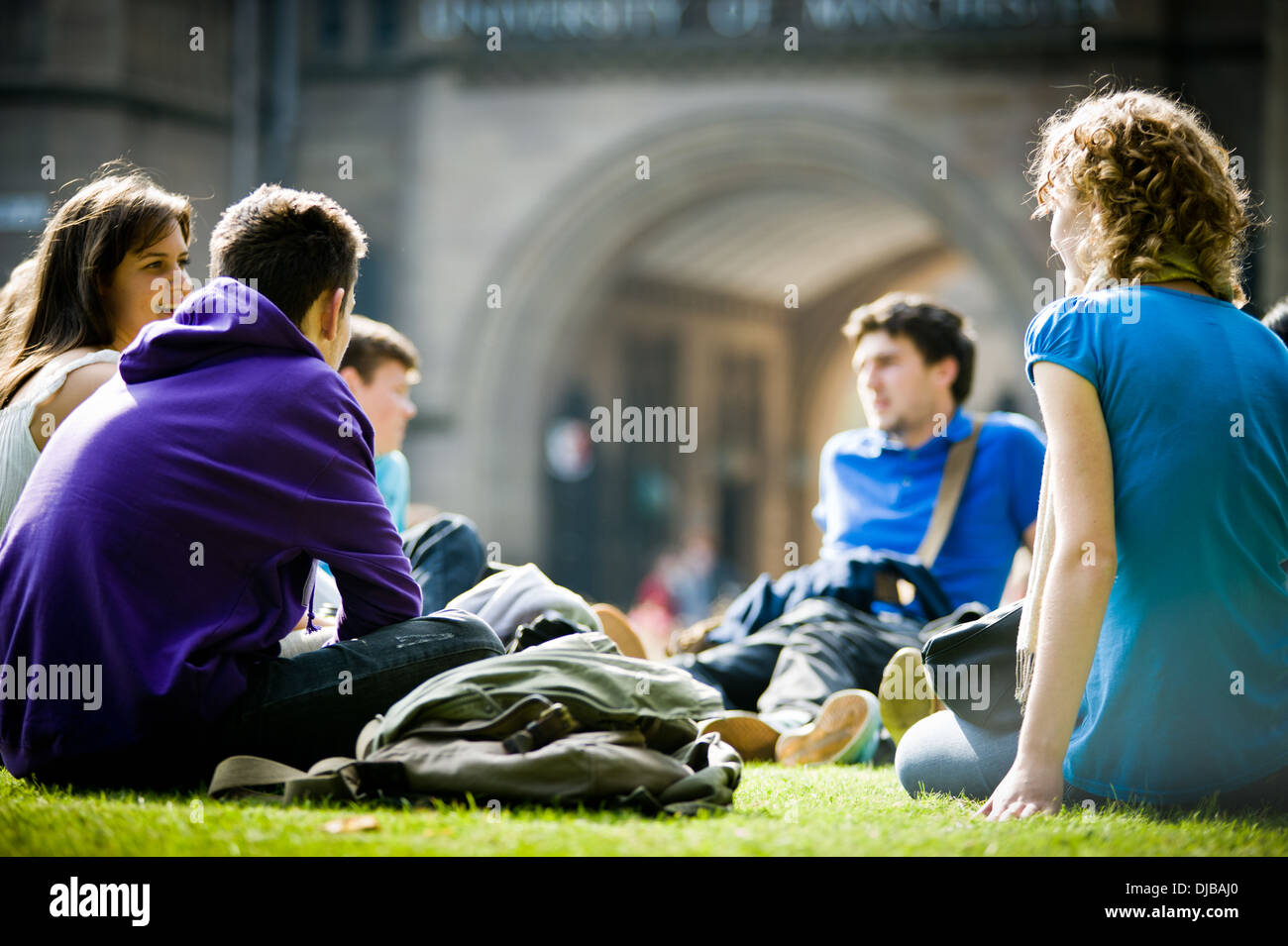 Gruppe von Multi Rasse Manchester Studenten saßen auf Rasen auf dem Campus im Sonnenschein reden und lachen, schauen glücklich Stockfoto