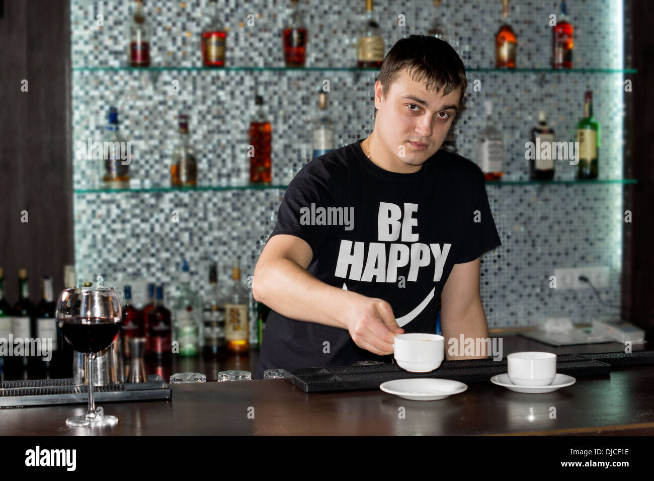 Junge kaukasischen Barkeeper mit einem lustigen T-shirt mit einer optimistischen Botschaft mit Kaffee an der Bar mit Regalen mit Flaschen von alkoholischen Getränken in den Hintergrund. Stockfoto