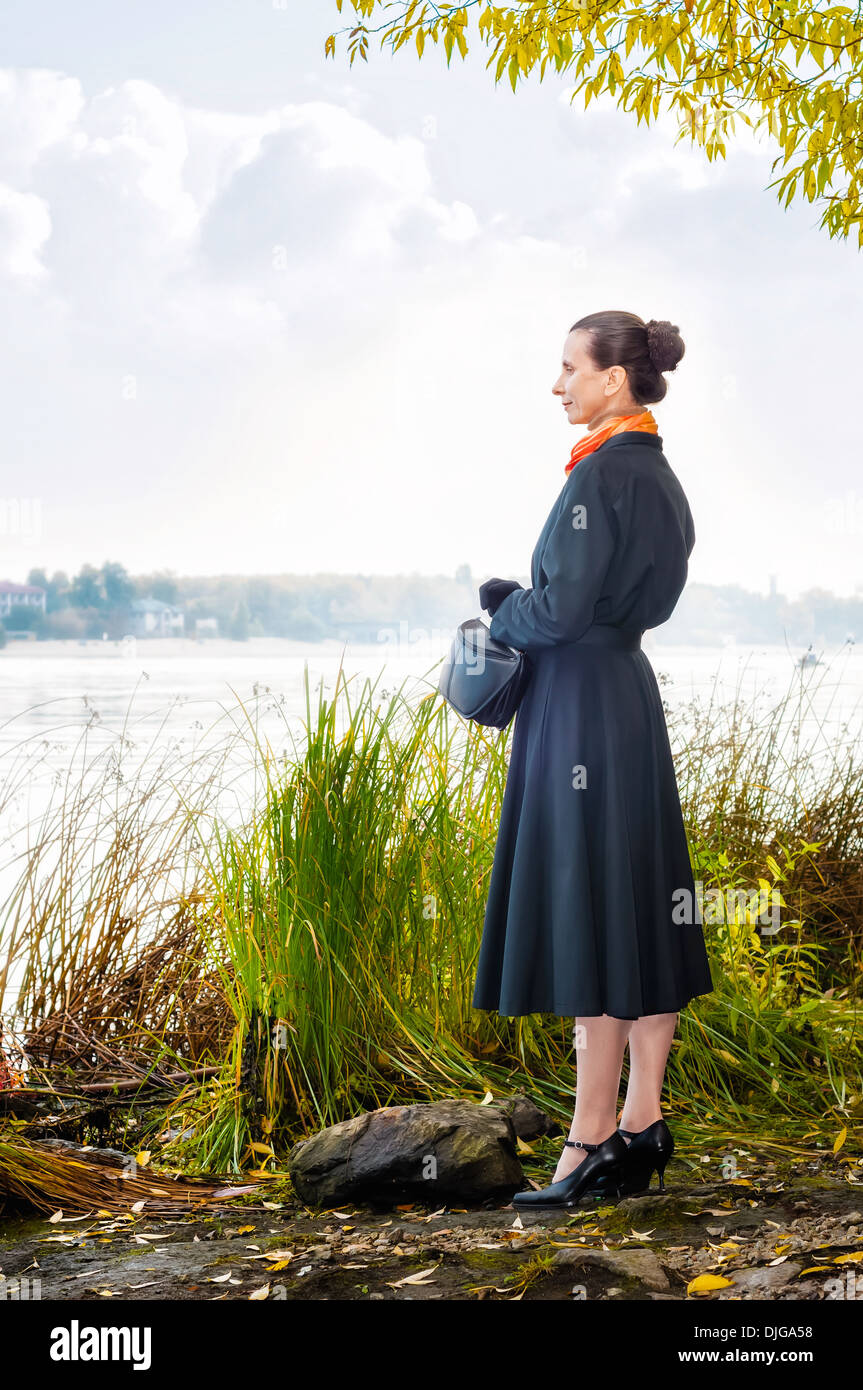 Elegante senior Business-Frau mit einer Tasche und einem orangen Schal, zu Fuß entlang des Flusses, unter den Bäumen im Herbst Stockfoto