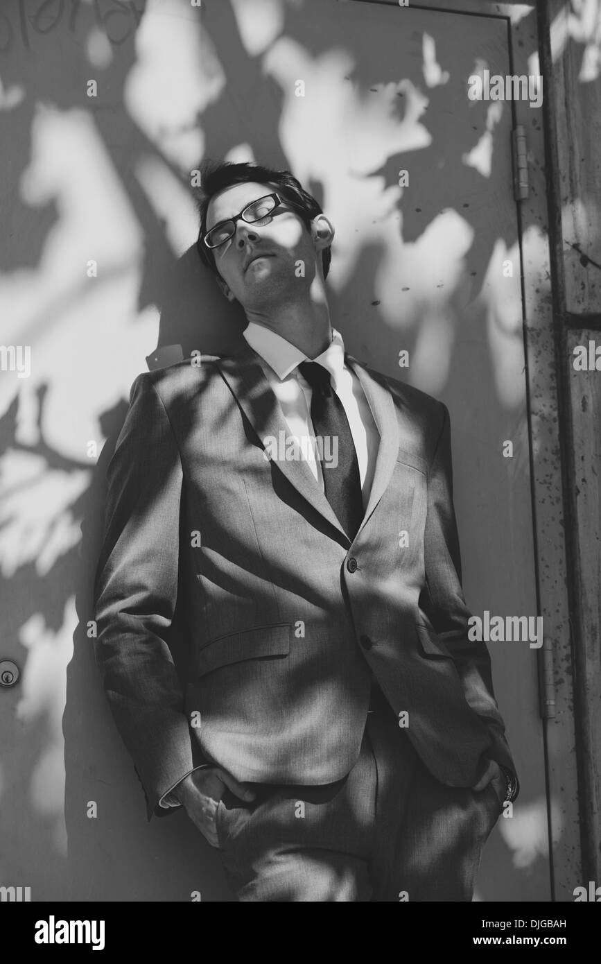 Ein Business-Mann, gekleidet in Anzug und Krawatte, gelehnt eine Tür einen Schatten mit seinen Augen geschlossen und die Hände in der Tasche. Stockfoto