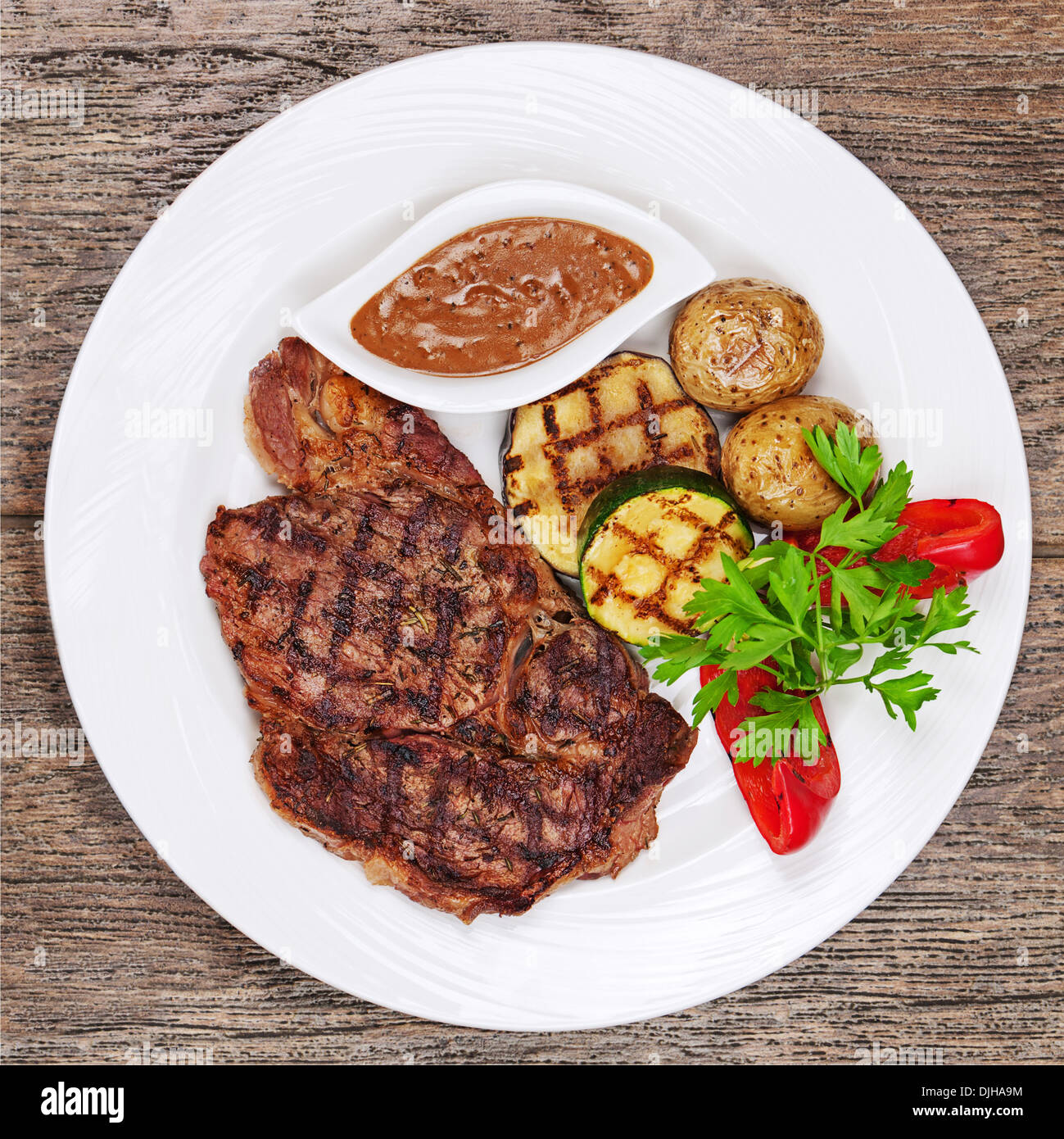 Gegrillte Steaks, gebackene Kartoffeln und Gemüse auf weißen Teller auf hölzernen Hintergrund. Stockfoto