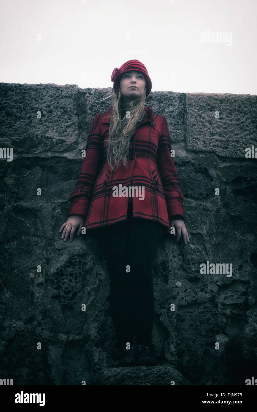 eine Mädchen in einem roten Mantel mit Hut ist an einer alten Steinmauer gelehnt. Stockfoto