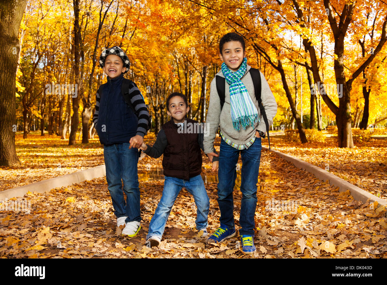 Drei glückliche, lachende schwarze Jungen , Brüder 5-10 Jahre zusammen gehen Hand in Hand im Park tragen Rucksäcke und Herbst Kleidung in Maple Park Stockfoto