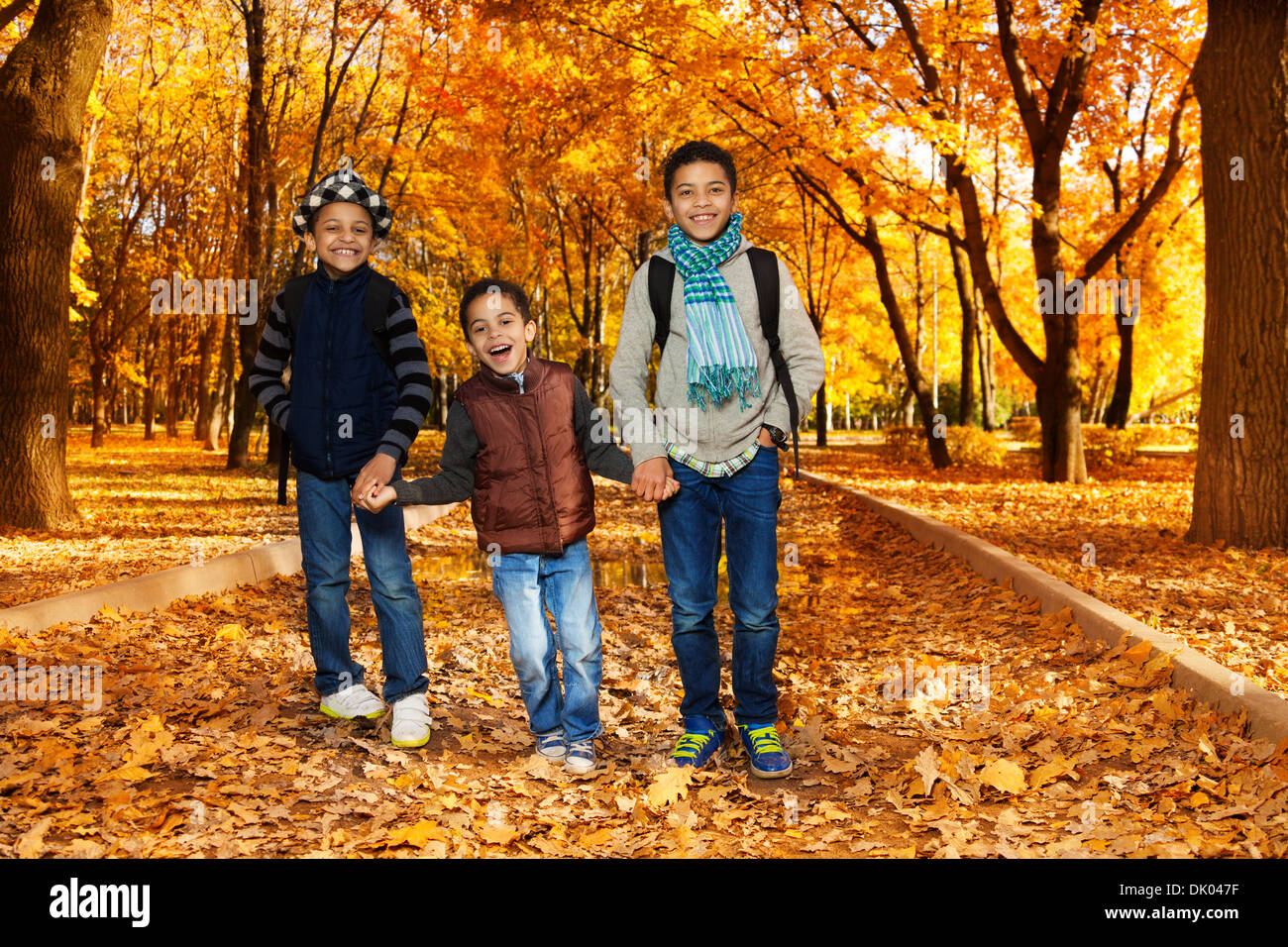 Drei glückliche, lachende schwarze Jungen , Brüder 5-10 Jahre zusammen gehen Hand in Hand im Park tragen Rucksäcke und Herbst Kleidung in Ahorn und Eiche Park mit vielen Herbst rot und orange Blätter Stockfoto