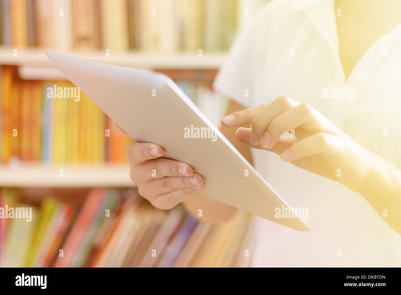 Weibliche Hände halten Tablet-Computer, Bücherregal im Hintergrund - Bibliothek oder College-Klasse oder Krankenhaus Innenraum, warme Sonnenlicht. Stockfoto