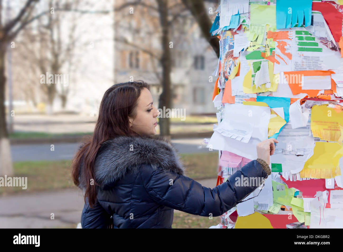 Junge Frau liest eine Mitteilung über einen öffentlichen Aushang im freien bedeckt mit bunten Noten und Inserate Stockfoto