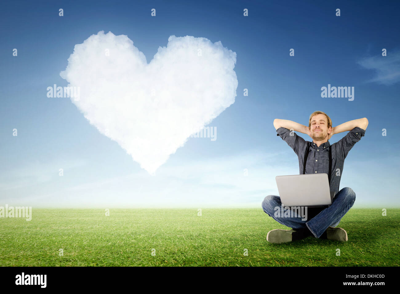 ein Mann sitzt mit Laptop entspannt auf der Wiese mit einer Wolke in Herzform im Hintergrund Stockfoto