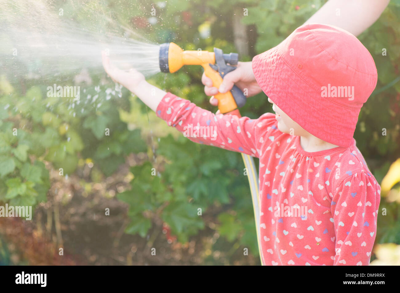 Lifestyle Sommer-Szene. Kleine Mädchen spielen mit Eltern im Garten, Gefühl Wasser aus Sprinkleranlagen Stockfoto