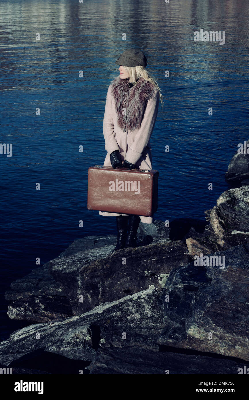 eine Mädchen in einem rosa Mantel steht am Meer auf den Steinen mit einem Koffer Stockfoto
