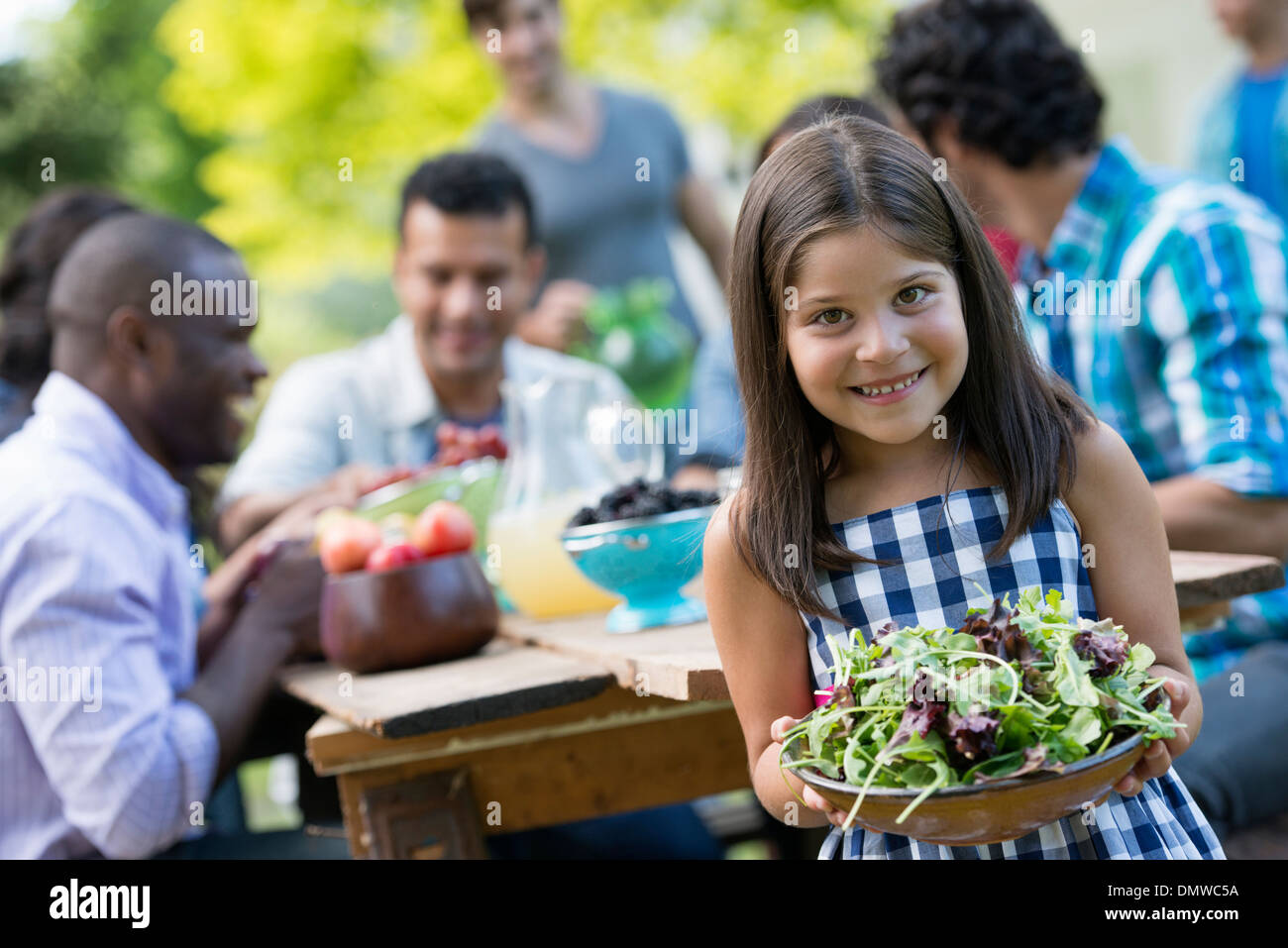 Erwachsene und Kinder an einem Tisch im Garten. Ein Kind hält eine Schüssel mit Salat. Stockfoto