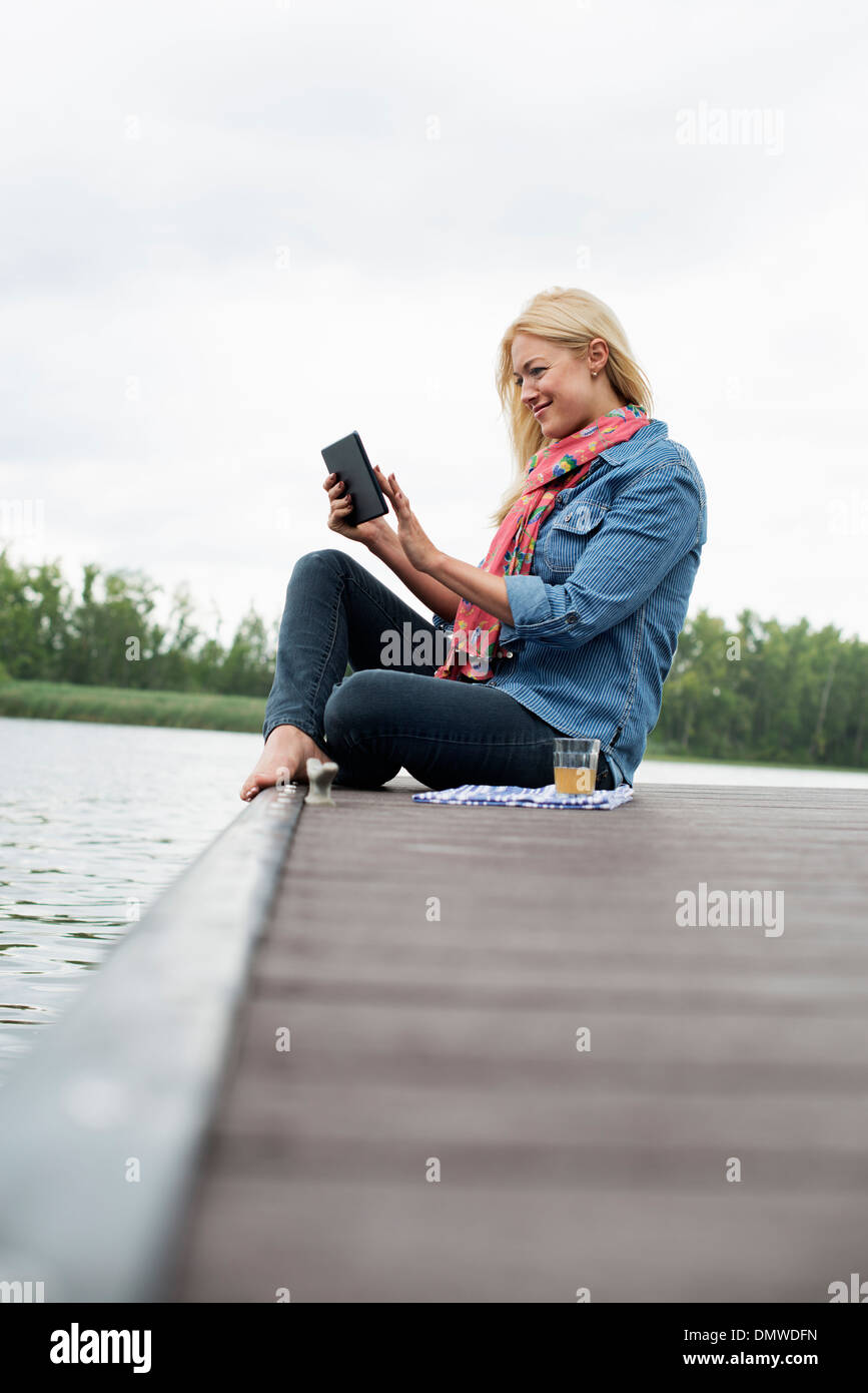 Eine Frau sitzt auf einem Steg an einem See mit einem digitalen Tablet. Stockfoto